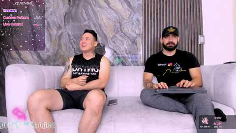 Media: Video of two men, one in a black tank top, shorts, and headphones, the other in a black cap, t-shirt, and jeans, seated on a white couch, with a virtual background featuring a galaxy and chat window.