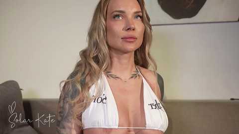 Media: A video of a fit, blonde woman with tattoos, wearing a white halter bikini top. She has a stern expression and stands against a beige wall with minimalistic art in the background.