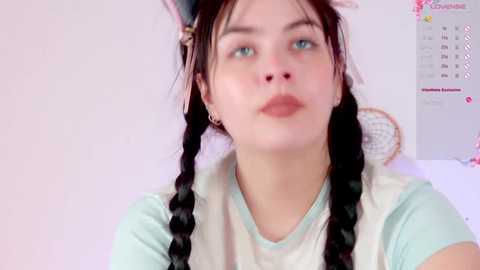 Media: Video of a young woman with fair skin, blue eyes, and long dark hair in braids, wearing a light blue shirt and a white apron. She has a serious expression. Background shows a calendar and a white wall.