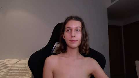 Media: Video of a young, slender, shirtless white man with shoulder-length brown hair, sitting in a black gaming chair in a dimly lit room with beige walls and a partially visible beige couch.