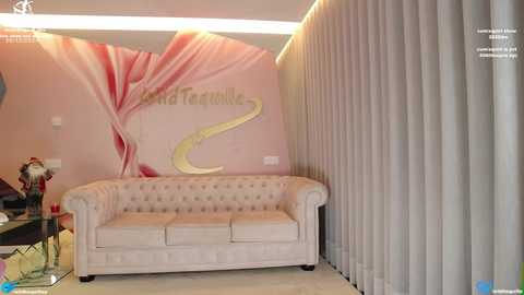 Media: Video of a modern, pastel-colored living room featuring a cream-colored tufted sofa, beige carpet, pink and white \"Ritz Carlton\" logo wall art, and tall white vertical blinds.