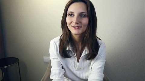 Media: Video of a young Caucasian woman with straight, medium-length brown hair, wearing a white button-up shirt, seated indoors against a plain beige wall, smiling softly.