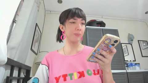 Media: A video of a young woman with short black hair, wearing pink earrings and a pink t-shirt, taking a selfie in a brightly lit room with framed art and a window in the background.