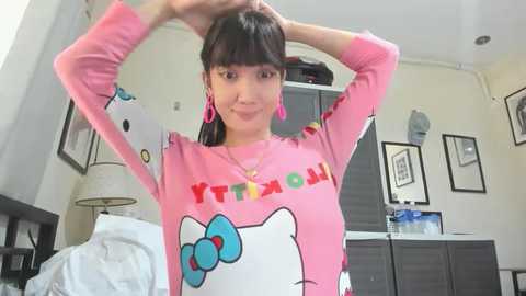 Media: Video of a young woman with straight black hair, wearing a pink Hello Kitty sweatshirt, pink earrings, and standing in a messy bedroom with framed pictures and a nightstand.