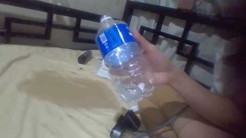 Media: A video showing a person pouring water from a blue plastic bottle onto a beige bedsheet, with a black metal bedframe in the background.