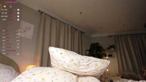 Media: Video of a dimly lit bedroom with white floral-patterned bedding, beige curtains, and a small plant on a shelf in the background.