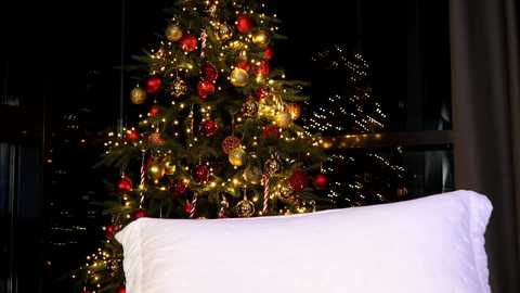 Media: A video of a Christmas tree adorned with red, gold, and white ornaments, nestled against a dark window, with a white pillow in the foreground.