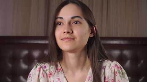 Media: Video of a young woman with long, straight brown hair, fair skin, and a slight smile, wearing a floral-patterned blouse, seated against a dark brown, tufted leather headboard.