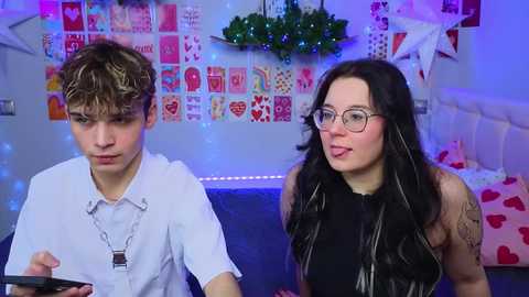 Media: Video of a young, pale-skinned, androgynous boy in a white shirt and a young, fair-skinned woman with long black hair and glasses, both seated in a cozy, heart-decorated room.