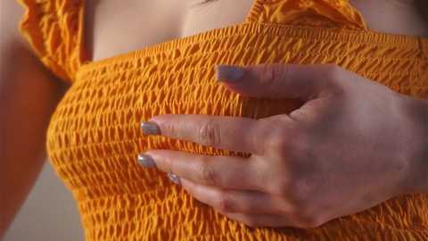 A close-up video of a woman with light skin, wearing a bright orange, textured, short-sleeved top. Her left hand, with glittery blue nail polish, is placed gently on her chest. The background is blurred.