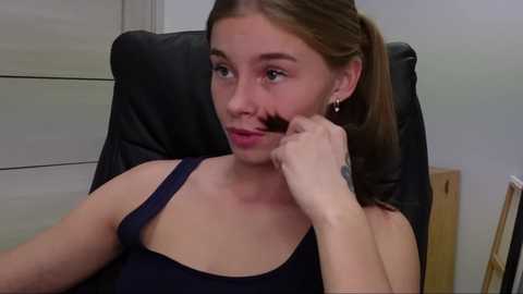 Media: A video of a young woman with light skin and brown hair in a ponytail, wearing a dark blue tank top, sitting in a black leather office chair. She holds a small black object near her mouth.