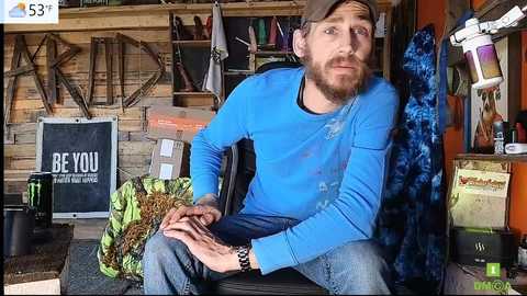 Media: Video of a bearded man with a light beard, wearing a blue shirt, jeans, and a baseball cap, sitting on a wooden chair in a rustic, cluttered workshop.