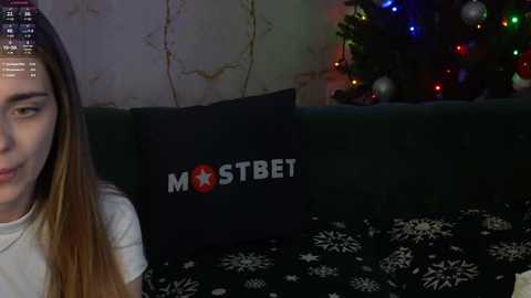 Media: Video of a young woman with long brown hair, wearing a white shirt, sitting on a dark couch with a black pillow featuring a red star and white text \"MOSTBET.\" Christmas tree with colorful lights in the background.