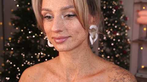 Media: Video of a fair-skinned, freckled woman with blonde hair wearing white, heart-shaped earrings. She has a confident smile, and the background features a decorated Christmas tree with warm, bokeh lights.