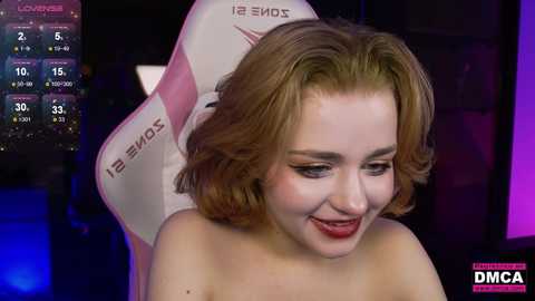 Media: Video of a fair-skinned, curly-haired woman with light makeup, smiling. She's sitting on a pink and white gaming chair. Background shows a dimly lit room with a calendar and clock. \"DMM\" logo at bottom right.