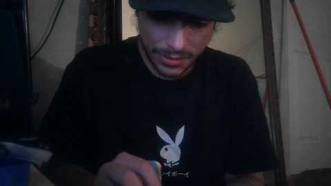 Media: A dimly lit video of a young man in a black Playboy T-shirt, gray cap, and mustache, looking down, in a cluttered, dimly lit room with cables and a blue object in the background.