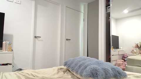 Media: Video of a modern, minimalist bedroom with white walls, a white bed, a blue fur pillow, and a large mirror reflecting a white dresser and TV.