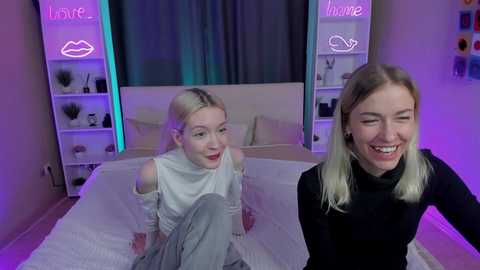 Video of a smiling, blonde woman in a black turtleneck, sitting on a bed with white sheets, in a modern, softly lit bedroom with purple accents.