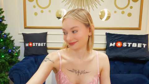 Video of a smiling blonde woman in pink lingerie, with tattoos on her chest and arms, seated on a blue couch.