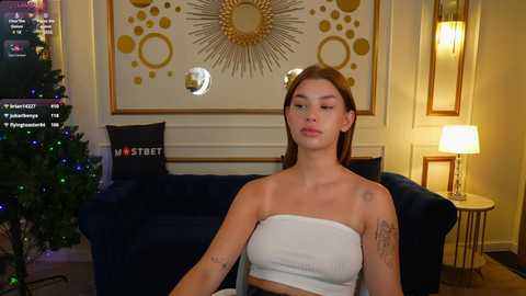 Media: Video of a young woman with straight, shoulder-length brown hair, wearing a white tube top, sitting on a dark blue couch in a warmly lit room with a Christmas tree and a gold sunburst wall art.