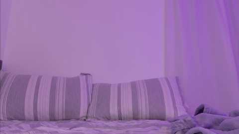 Media: A video of a minimalist bedroom with a white wall, purple lighting, and a bed with striped pillows in shades of white and gray. The bedspread is white with subtle patterns.