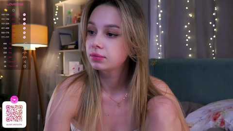 Media: A video of a young woman with long blonde hair, fair skin, and a contemplative expression, sitting on a bed in a dimly lit bedroom adorned with fairy lights and a lamp.