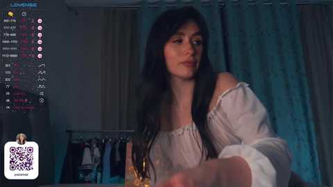 Media: Video of a young woman with long black hair, fair skin, and a slim physique, wearing a white off-shoulder blouse, standing in a dimly lit room with a closet full of clothes in the background.