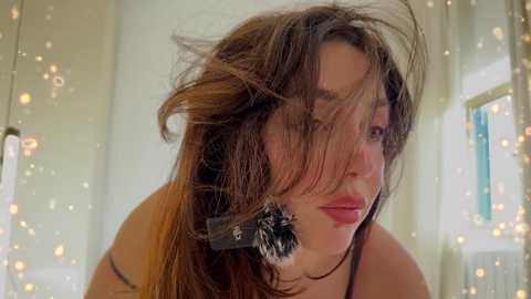 Media: Video of a woman with messy, voluminous brown hair, wearing a black headband and a white top, in a softly lit room with string lights and a window.