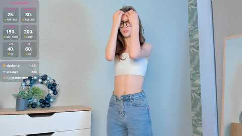 Media: A young woman with glasses, wearing a white crop top and high-waisted jeans, stands in front of a wall displaying weather and news updates.