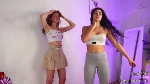 Media: Video of two women in a room. One with light skin and long hair wears a white crop top and beige skirt. The other, with darker skin and long black hair, wears a white crop top and gray leggings.