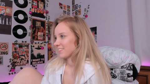 Media: Video of a smiling, blonde woman in a white shirt, sitting on a bed with a white duvet and a cluttered wall covered in posters and records.