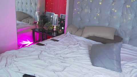 Media: Video of a modern, dimly lit bedroom with white bedding, a tufted headboard, and a red accent wall. Decorative string lights adorn the headboard, and a bedside table holds a lamp and a vase with flowers.