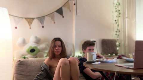 Media: Video of a young woman with medium-length brown hair, wearing a gray shirt, sitting on a beige sofa, while a young man with short brown hair, dressed in a red shirt, plays a video game on a table in a cozy, dimly lit living room.