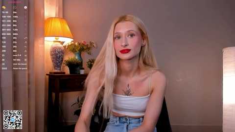 Video of a blonde woman with long hair, red lipstick, wearing a white crop top and high-waisted jeans, sitting in a softly lit room with a lamp and potted plant in the background.