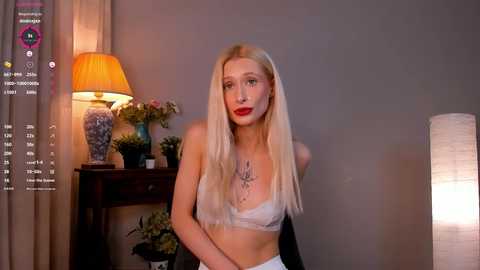 Video of a blonde woman with fair skin, red lipstick, wearing a white bra, sitting in a room with a table lamp, plants, and a gray wall.