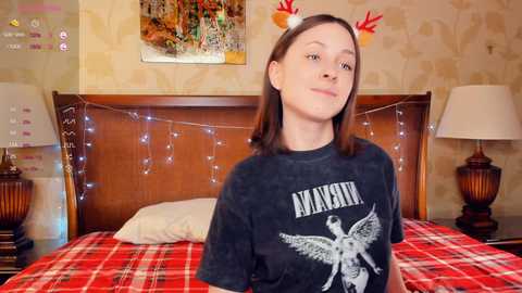 Media: Video of a young Caucasian woman with shoulder-length brown hair, wearing a black Metallica t-shirt and red reindeer antlers, sitting on a red plaid bed with wooden headboard and string lights, in a cozy, patterned wallpaper bedroom.