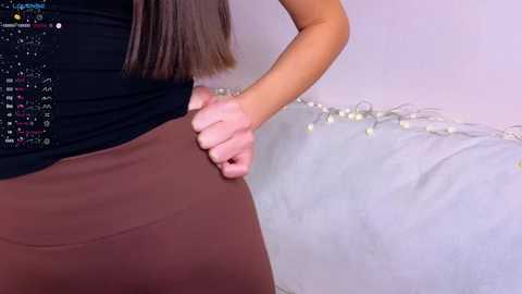 Media: A video of a woman in a black top and brown skirt, adjusting a light blue cushion with string lights on a beige wall.