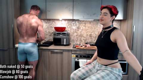 Media: Video of two people in a modern kitchen: a shirtless man in tight blue shorts and a woman in a black crop top and plaid pajama pants.