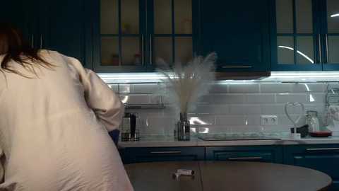 Media: Video of a woman in a beige robe, standing in a sleek, modern kitchen with dark blue cabinets, white subway tiles, and a stainless steel sink.