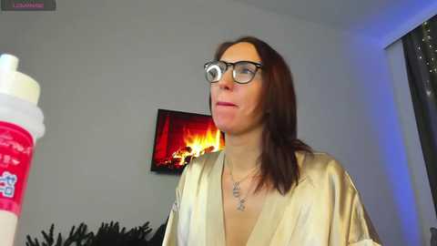 Media: Video of a middle-aged Caucasian woman with straight brown hair, wearing glasses and a silky gold robe, holding a coffee cup, standing in a modern living room with a lit fireplace and dark curtains.