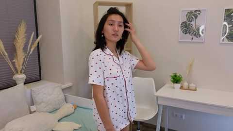 Media: Video of an Asian woman with medium-length black hair, wearing a white short-sleeved shirt with black polka dots, standing in a minimalist, modern room with white furniture and greenery.