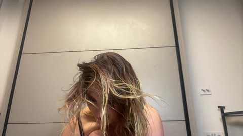 Media: A video of a partially obscured woman with long, wet, blonde-streaked hair, standing in a modern, minimalist bathroom with white tiled walls and a black metal towel rack.