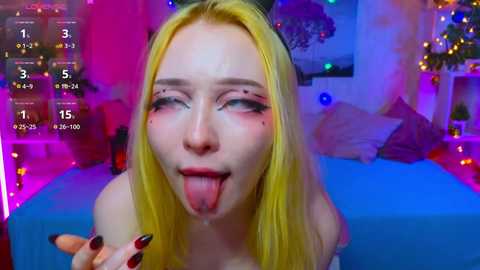 Media: A video of a young, fair-skinned woman with long, straight blonde hair, wearing heavy makeup, sticking out her tongue, and holding a finger. Background shows a colorful, lit room with festive decorations.