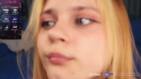 Media: Video of a young woman with long blonde hair, fair skin, and pink makeup, looking contemplative. In the background, a live stream interface with weather data and \"Loud and Clear\" text.