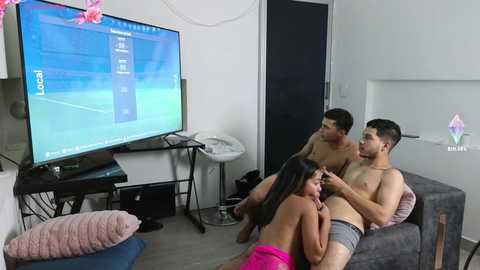 Media: Video of a young, fit, shirtless man and a topless woman, both with medium skin tones, sitting on a sofa in a modern living room. A large TV displays a tennis game on a white wall.