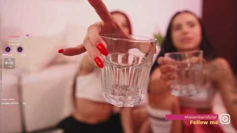 Video of a woman with red nails pouring water into a glass, with another woman in the background, both in casual attire.