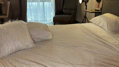 Media: Video of a cozy bedroom with a neatly made bed featuring white sheets and fluffy pillows. In the background, a window with sheer white curtains allows soft natural light to filter in. A brown leather armchair and a small wooden table with a lamp are positioned near the window.