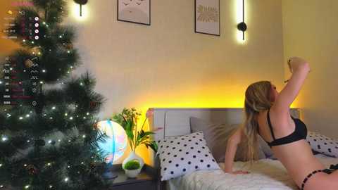 Media: Video of a blonde woman in black lingerie dancing in a bedroom with a lit Christmas tree, bed with white polka-dotted sheets, and modern wall art.