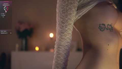 Media: Video of a woman's upper back, wearing a lace top, showing a tattoo of Hello Kitty and a cat on her left side. Background features dimly lit candles and a vase with flowers.