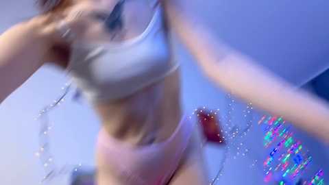 Media: Video of a light-skinned woman with long brown hair, wearing a white bra and pink panties, dancing in a dimly lit room with a blue glow.